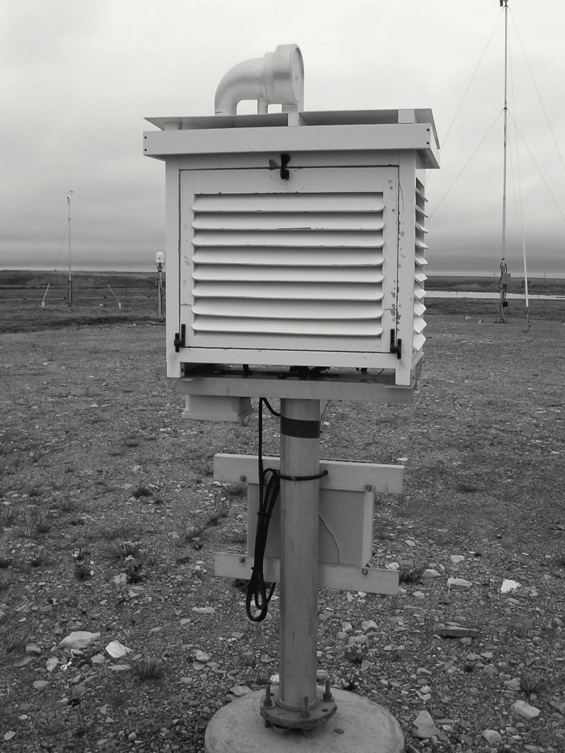 Un abri météorologique en bois blanc, monté sur un pied à hauteur de poitrine au milieu d'un terrain de graviers : ses parois sont faites de lames inclinées qui laissent passer l'air et arrêtent le soleil.