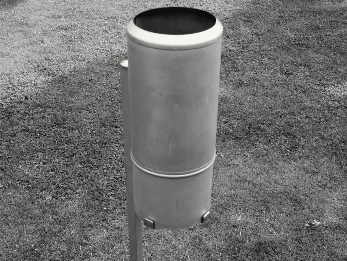Un tube de métal gris posé sur un piquet planté dans une pelouse, ouvert vers le ciel : c'est un pluviomètre, et l'eau qui tombe entre par le haut.