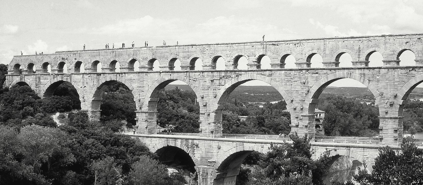 Un aqueduc romain de pierre à trois étages d'arcades enjambe une vallée boisée ; l'étage du haut est un long rang de petites arches, les deux étages du bas portent de grandes arches.